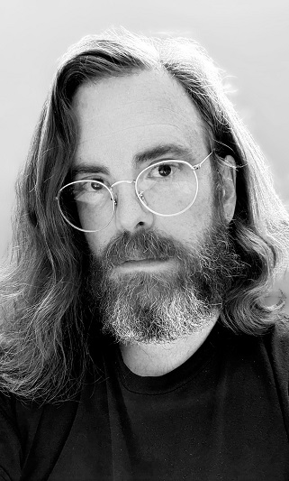 A portrait of Julian Day in front of a neutral background. He is wearing a dark t-shirt and gold-rimmed glasses.