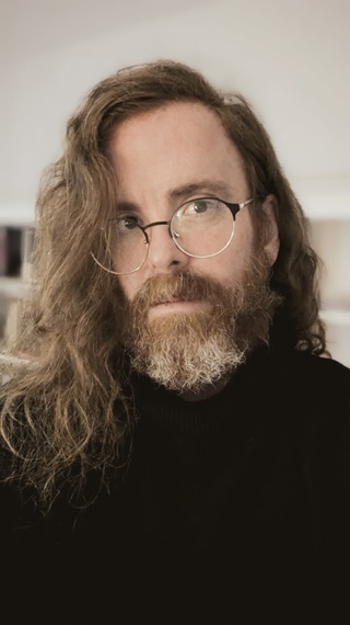 A portrait of Julian Day. He is wearing a black turtleneck sweater, round black-and-gold glasses, and has long, wavy red hair and a greying red beard.